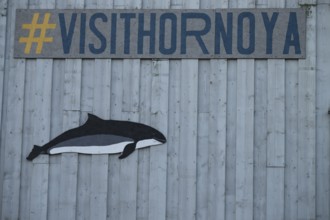 A whale image and a hashtag sign on a grey wooden wall with the slogan Visit Hornoya, Vardø,