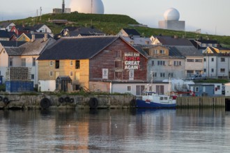 Houses and boats at a harbor against a green backdrop of hills in the evening light, Vardø,