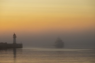 A lighthouse at the entrance to Vardö harbour at sunset with a ship in fog on calm water, Vardø,