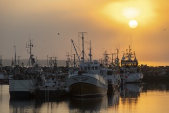 Fishing boats are moored in the harbor at sunset, the sea reflecting the warm colors of the sky,
