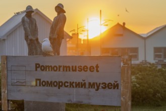 A seagull sits on a museum sign in Norwegian and Russian in front of sculptures and wooden