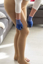 Woman puts on medical compression stockings with special gloves while standing in her home