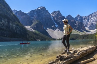 Female tourist standing on a log, admiring turquoise waters of moraine lake with canoers,