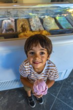 Young boy smiles while holding a scoop of strawberry ice cream in front of a display case filled