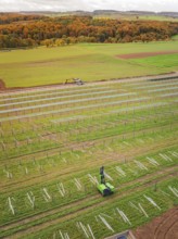 Tractor works in a green field with solar plant construction beginning, surrounded by autumnal