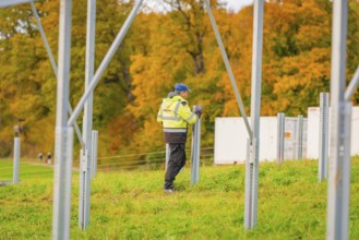 A worker wearing protective clothing erects metal structures on a grass-covered area in autumn,