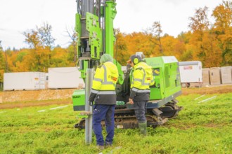 Two workers work with a green machine in front of autumn trees in a meadow, Energiewende,