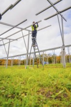 Man on a ladder assembles a metal structure on an open green area, Energiewende, construction of PV