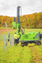 Two workers fix poles with a large, green machine in a field in an autumn landscape, Energiewende,