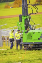 Two workers wearing safety clothing operate a green machine in a meadow in autumn, Energiewende,