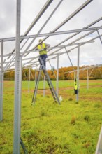 Workers stand on a ladder, assemble metal scaffolding over a green area surrounded by fields,