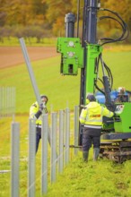 Workers install metal poles in a field with a large green machine in the background, Energiewende,
