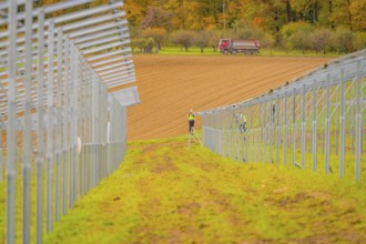 A farm worker on a ladder between metal structures with a tractor in the background in an autumn