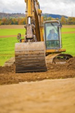 An excavator lifts soil on a field with a green landscape and a hilly background, Energiewende,