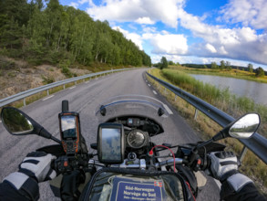 A motorcyclist's view of an empty country road surrounded by green trees and a lake under a blue