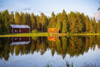 Two red houses are reflected in a quiet lake surrounded by forest under the warm light of the