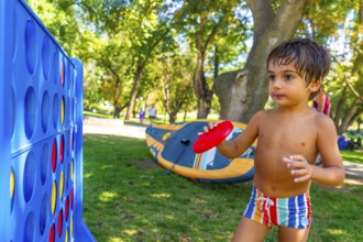 Young child holds a red flying disc, standing near a large connect four game in a park in kamloops,