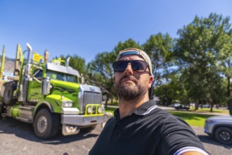 Trucker wearing sunglasses and baseball cap taking selfie with his logging truck in a parking lot