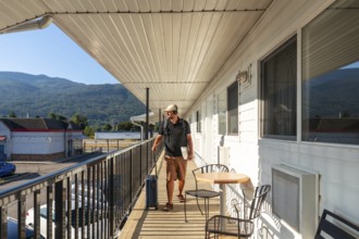 Male tourist wearing headphones and carrying a laptop strolls along a motel balcony, pulling