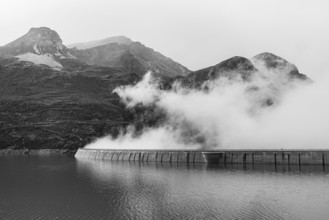 Fog rises over the dam of the Lac de Moiry reservoir, black and white photo, Val d'Anniviers,