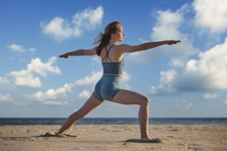 A woman practices Warrior 2 Virabhadrasana 2 pose on the beach during sunset, embodying strength