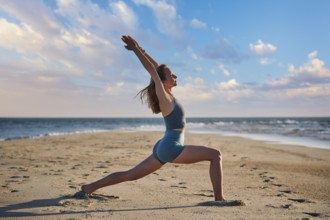 A woman performs Virabhadrasana 1, or Warrior 1 pose, on the beach during sunset. She stands strong