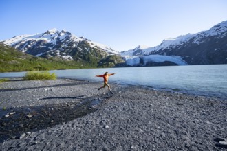 Young man jumping on a pebble beach on a glacial lake, Snowy Mountains and Portage Glacier on