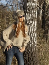 Slender pretty woman with fur hat and pompoms, red hair and warm winter colors between birch forest