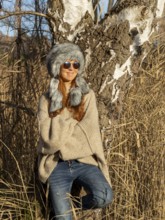 Slim woman with fur hat and sunglasses, red hair and warm winter colors between birch forest and