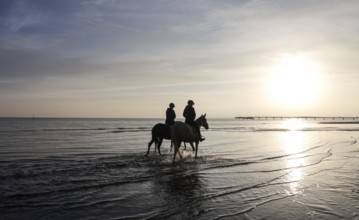 Two female riders ride their horses through the shallow water of the Baltic Sea at sunrise,