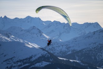 Paragliders flying over snowy mountain peaks in winter in evening light, Kitzbühel Alps, Tyrol,