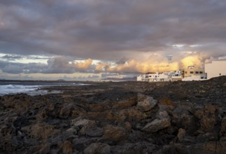 Black coast of volcanic rocks behind typical white houses of the village of La Santa, at sunset,