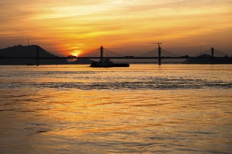 A boat sails on the river Brahmaputra during sunset, with the silhouette of a cable-stayed bridge