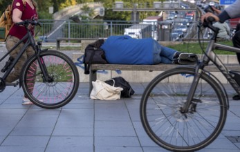 Man sleeping on a bench, travelling bag serves as pillow, in the city centre, passers-by walk by