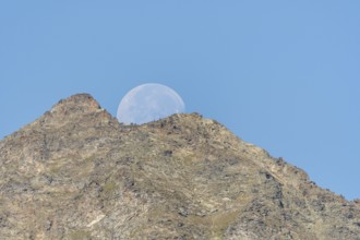 Majestic mountain peak stands tall against vibrant blue sky as large moon rises just behind it. The