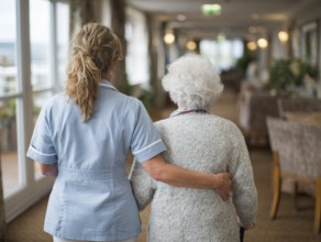 A carer assists an elderly woman walking along a bright corridor in a retirement home, nursing