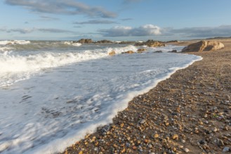 A wide sandy beach stretches out with scattered pebbles and beach shells. Gentle waves crash
