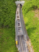 Aerial view of a railway tunnel stretching through green hills and a dense forest, construction of