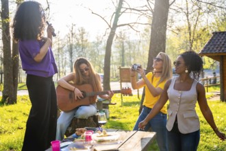 Four female friends enjoying a picnic in a park, singing, playing guitar, taking pictures and