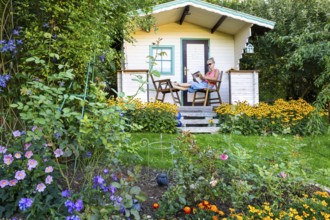 A woman sits relaxed on the small veranda of a garden shed and reads. Blooming summer flowers in