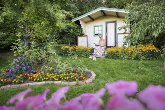 A woman in a dress sits relaxed on the small veranda of a garden shed and reads. Blooming summer