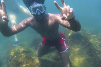 Young man snorkeling underwater, flashing the peace sign with both hands, enjoying the vibrant blue