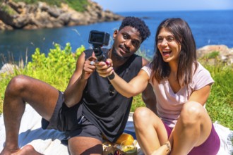 Happy young multi ethnic couple filming with action camera while having picnic by the sea during