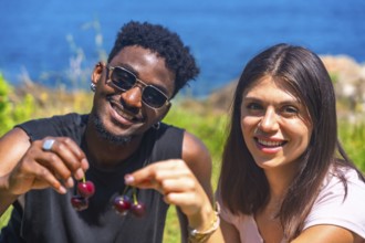 Interracial couple smiling while sharing fresh cherries, enjoying a sunny summer vacation by the