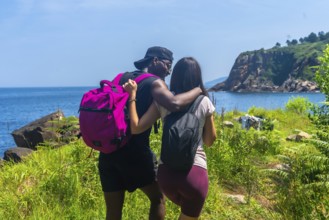 Interracial couple with backpacks walking on a path by the sea, embracing and enjoying the scenic