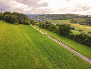 Car drives a straight road through wide green landscape with cloudy sky, Deer E- CarSharing,