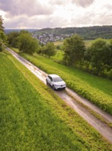Car drives on narrow road through green landscape with village on the horizon, Deer E- CarSharing,