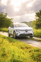 White car on a rural road with green meadows and cloudy sky in the background, Deer E- CarSharing,