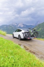 White car with bicycles on mountainous road, surrounded by green landscape and cloudy sky, electric