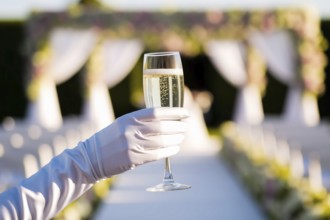 Close-up of a pristine white silk glove holding a champagne flute at a vibrant daytime wedding.